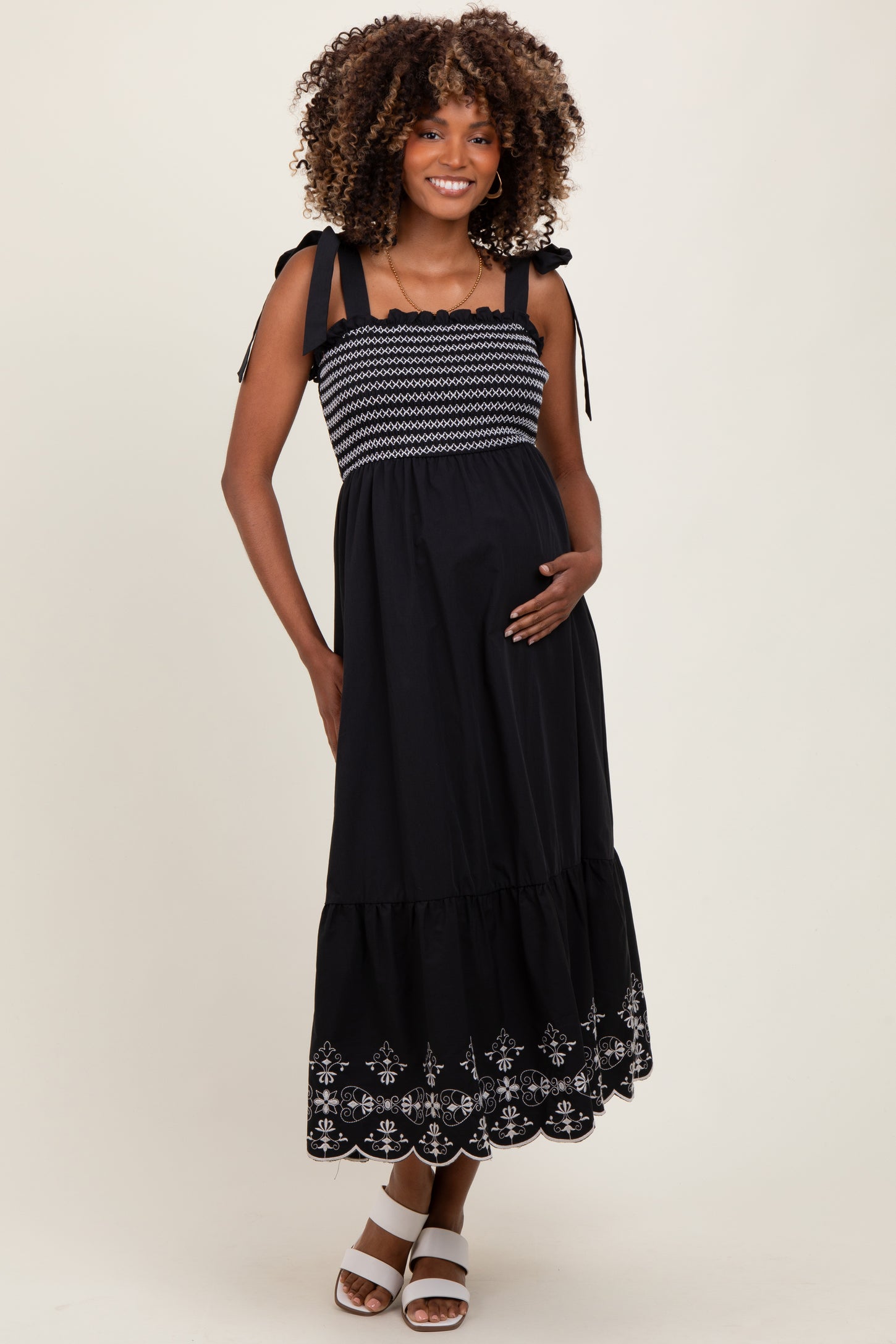 woman wearing a black maternity maxi dress with embroidered hem and smocked bodice, cradling her baby bump