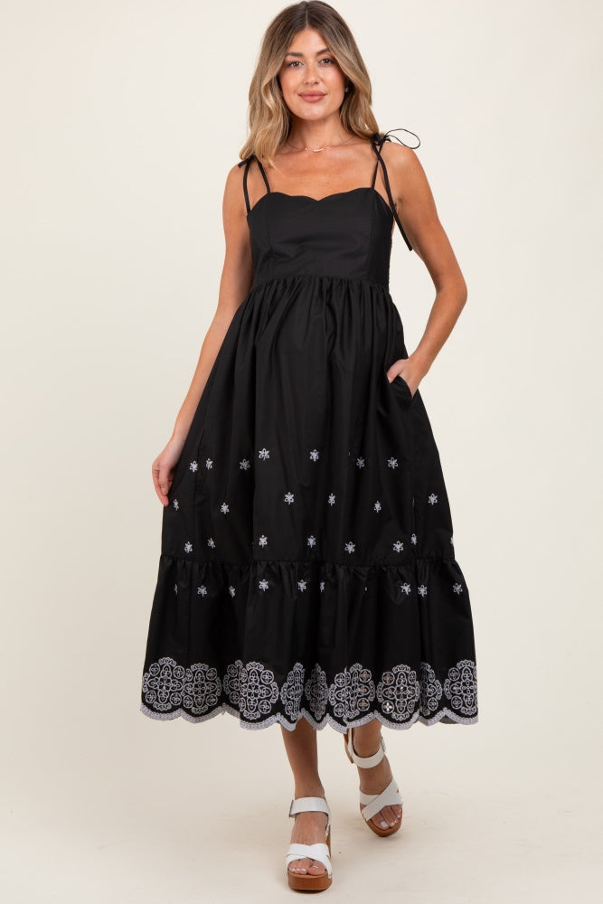 woman wearing a black maternity dress with floral embroidery and a tiered hem, featuring adjustable spaghetti straps