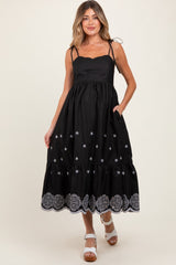 woman wearing a black maternity dress with floral embroidery and a tiered hem, featuring adjustable spaghetti straps