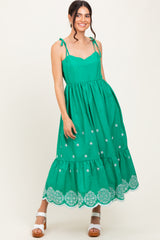 woman wearing a green sleeveless maxi dress with tie straps and floral embroidery along the hemline
