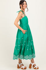 woman wearing a green maxi dress with embroidered details and tie straps, standing with pockets and white chunky heeled sandals