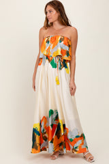 woman wearing a strapless floral maxi dress in orange, green, and yellow with a ruffled top and tie detail at the waist
