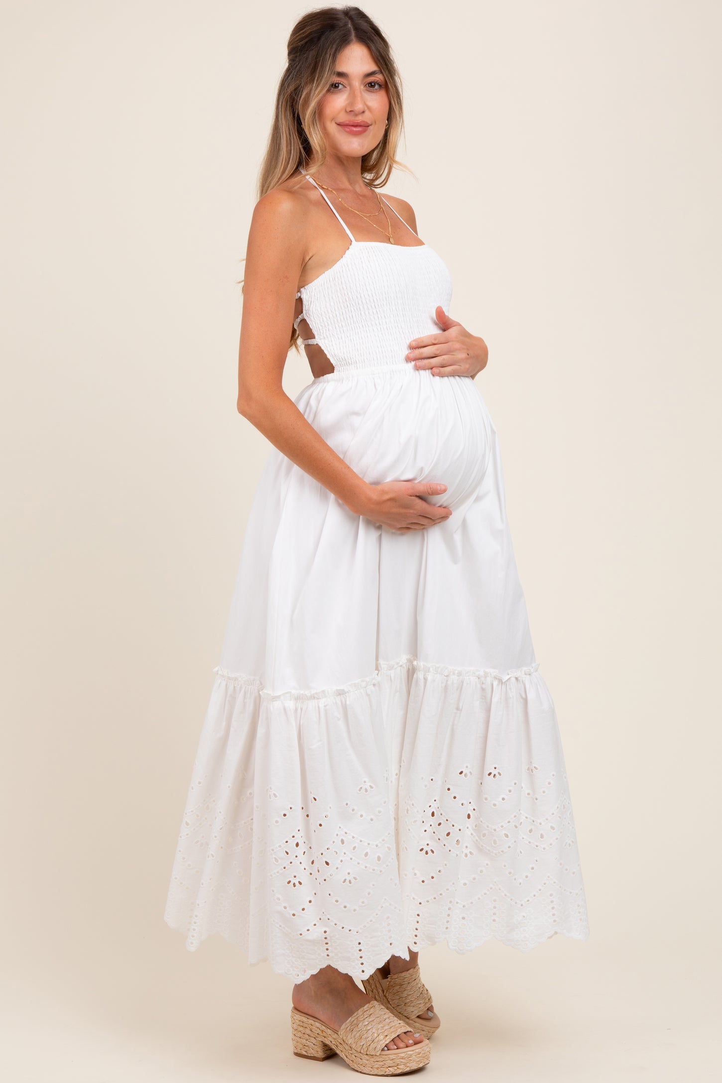 woman wearing a white maternity maxi dress with a smocked bodice and eyelet detailing, cradling her baby bump