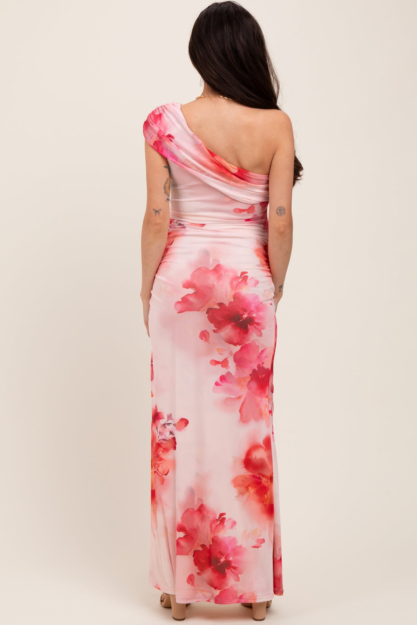 woman wearing a one-shoulder floral print maternity gown with pink and coral hues, showing her baby bump from the back