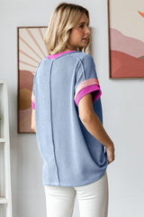 woman wearing a blue ribbed short sleeve top with pink striped details, showing the back view and styled with white pants