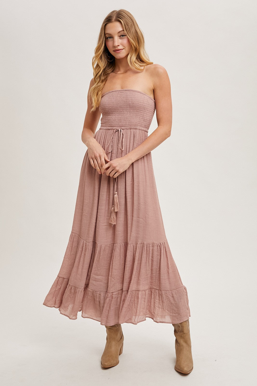 woman wearing a dusty pink strapless maxi dress with a smocked bodice and tiered hem, paired with tan ankle boots