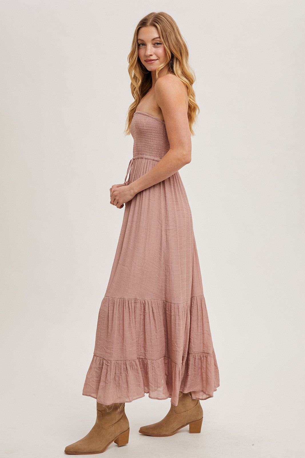 woman wearing a dusty rose strapless maxi dress with a smocked bodice and tiered ruffle hem, styled with tan ankle boots