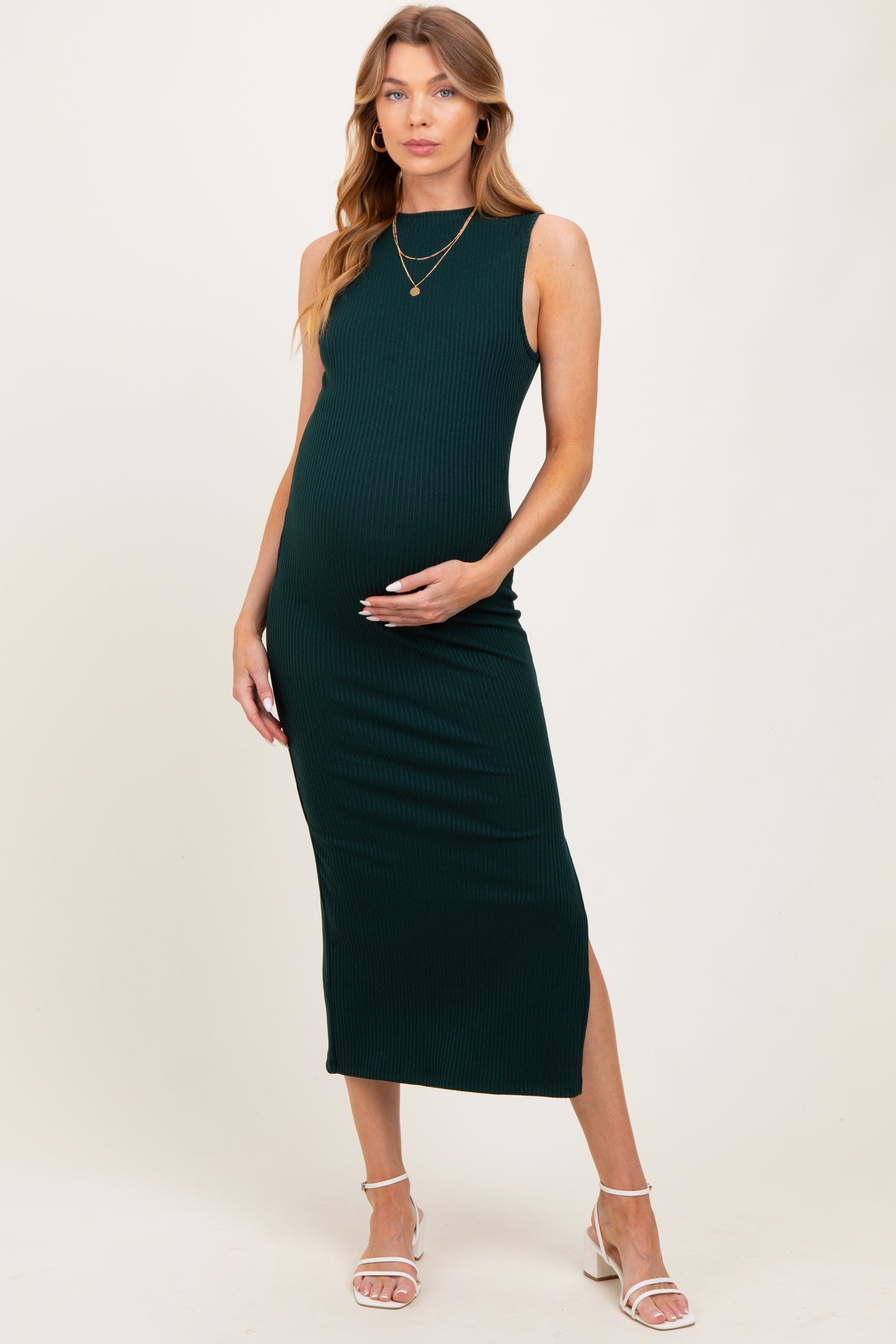woman wearing a dark green ribbed maternity dress, holding her baby bump, paired with strappy white sandals