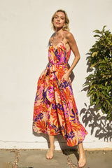woman wearing a vibrant floral maxi dress with spaghetti straps and a belted waist, standing next to a greenery backdrop