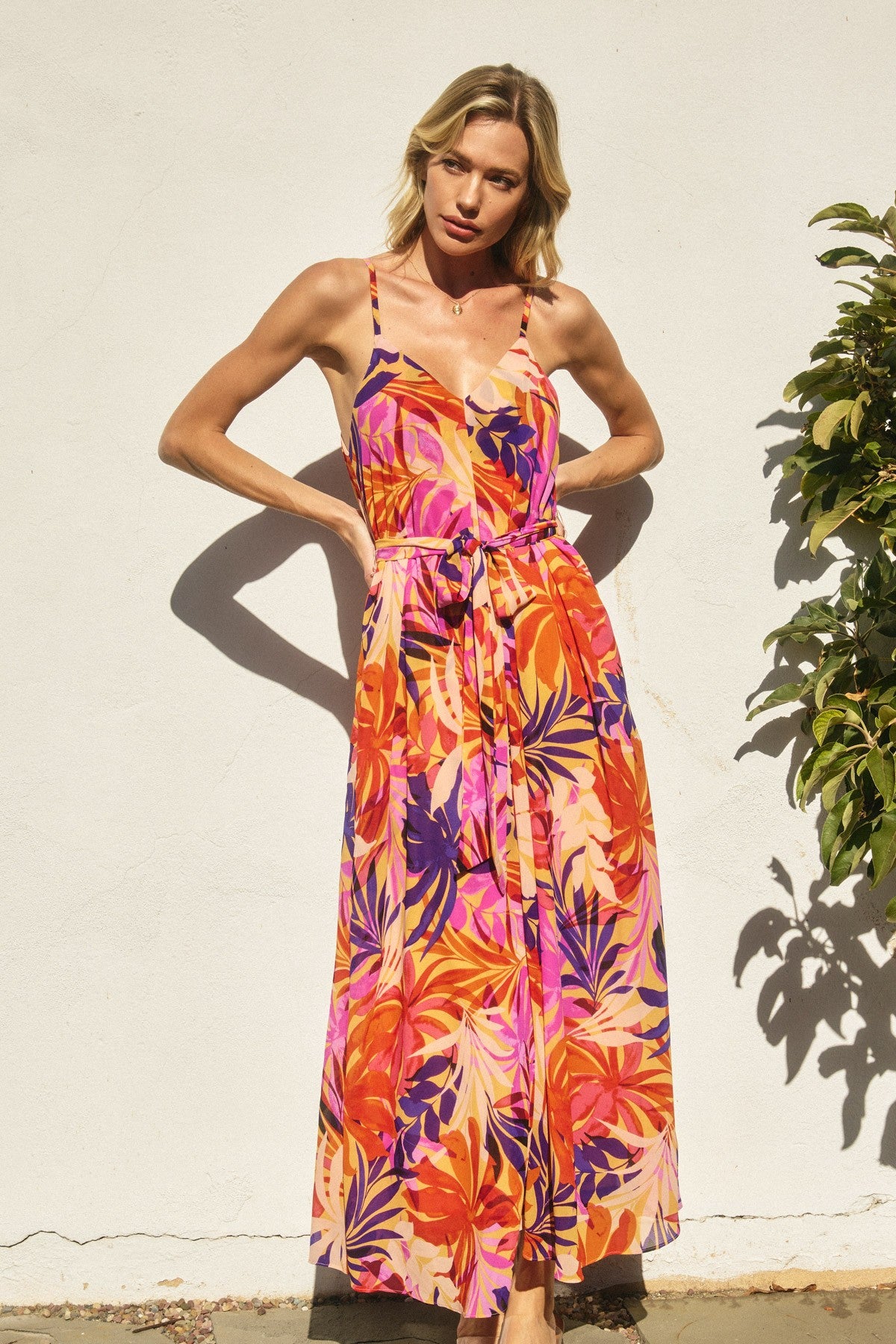 woman wearing a vibrant floral maxi dress with a tie waist and spaghetti straps, standing against a wall