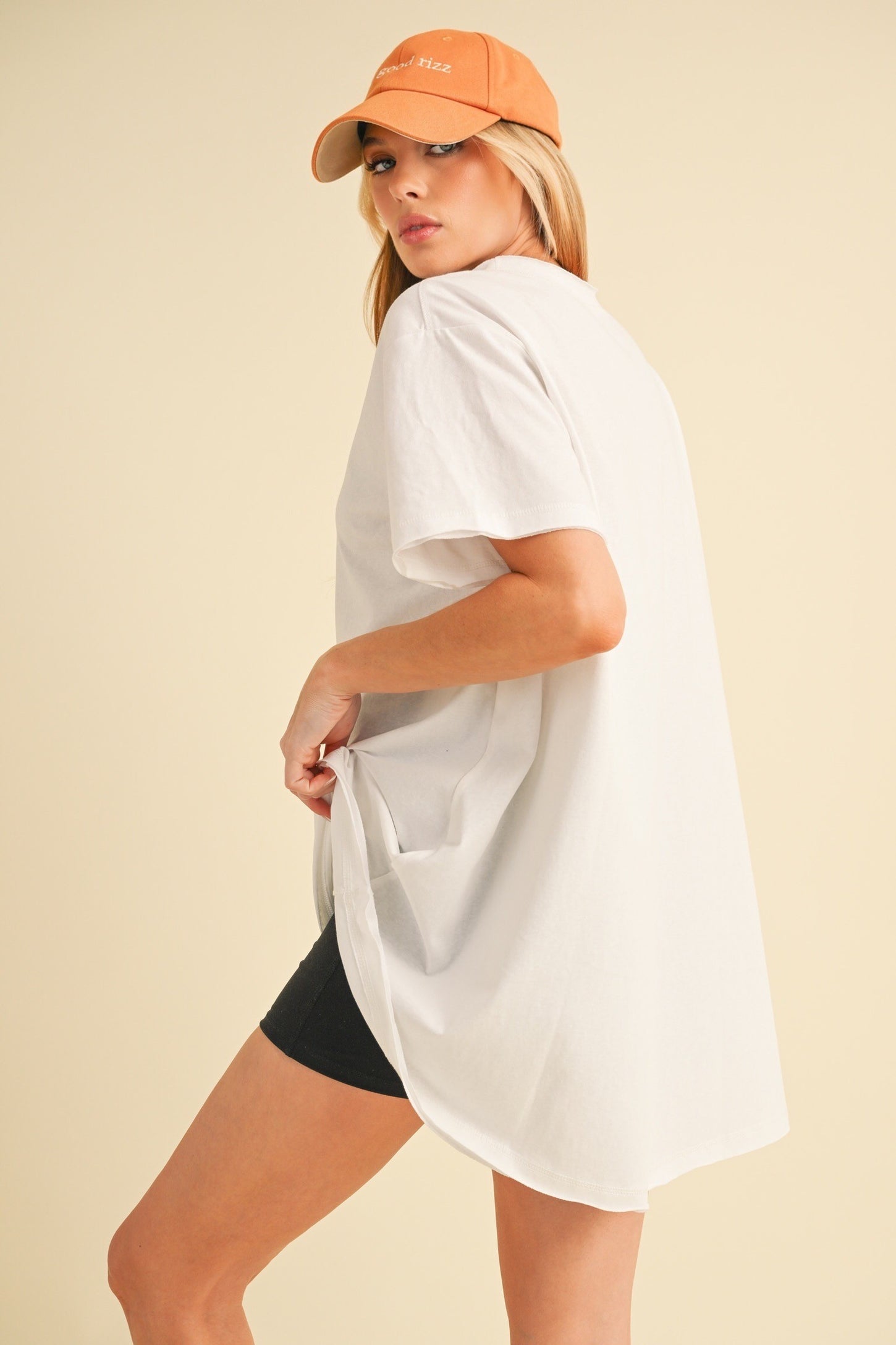 woman wearing an oversized white t-shirt and black shorts, turning to the side while wearing an orange cap