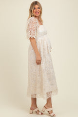 woman wearing a cream maternity dress with floral lace details and a baby bump, standing in profile