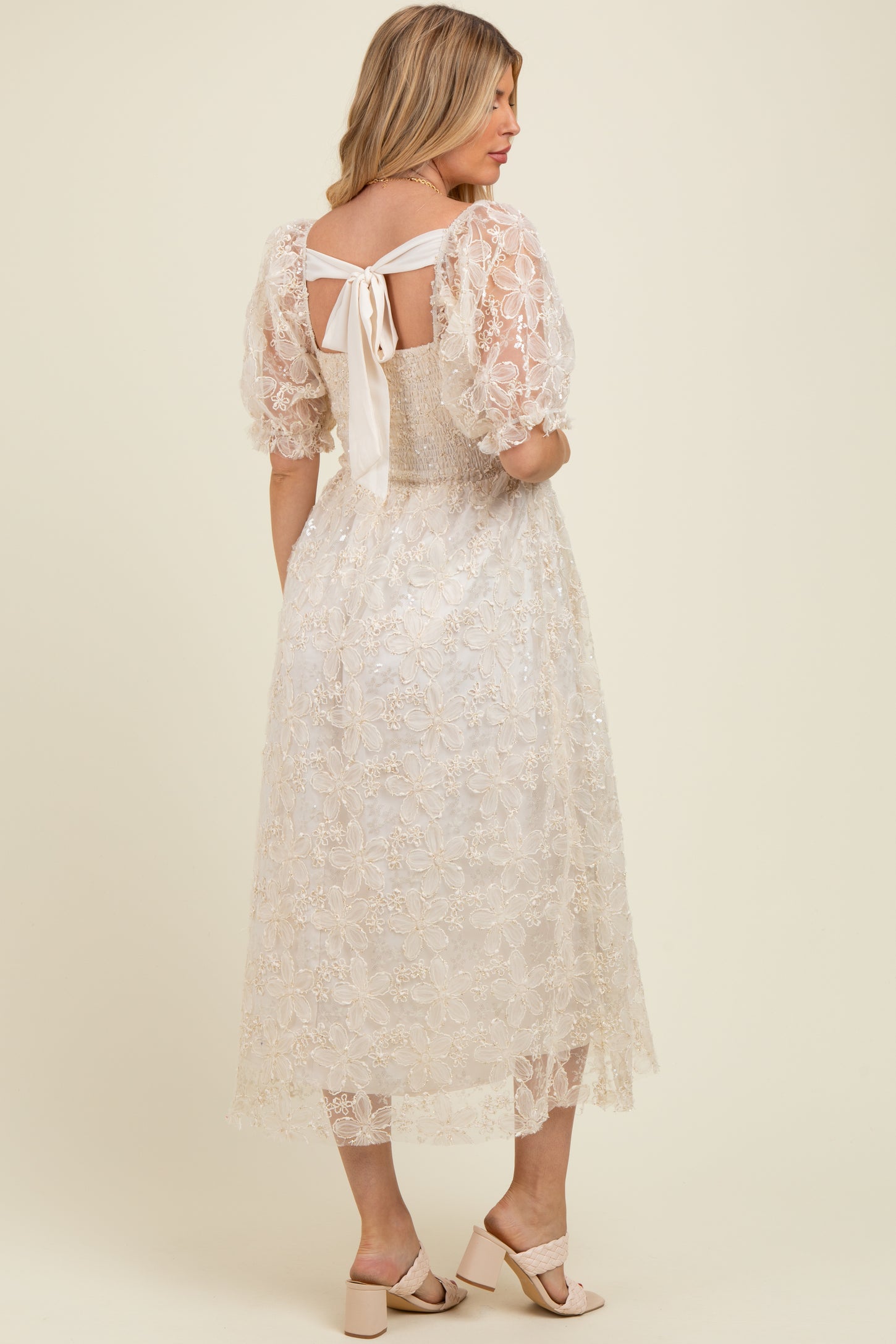 woman wearing a cream floral lace maternity dress with puff sleeves and a tied open back