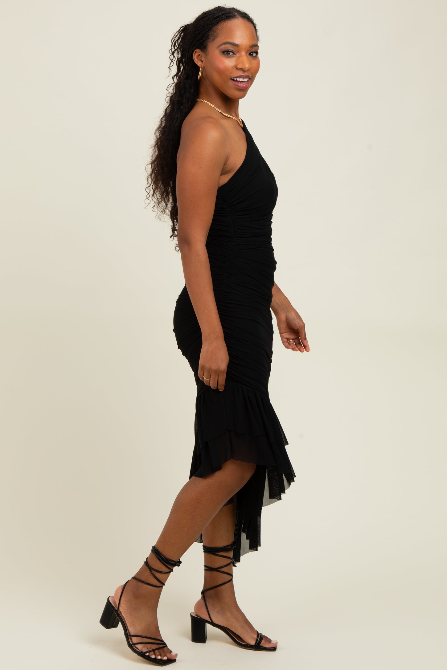 woman wearing a black one-shoulder ruched midi dress with a ruffled hem and strappy high-heeled sandals