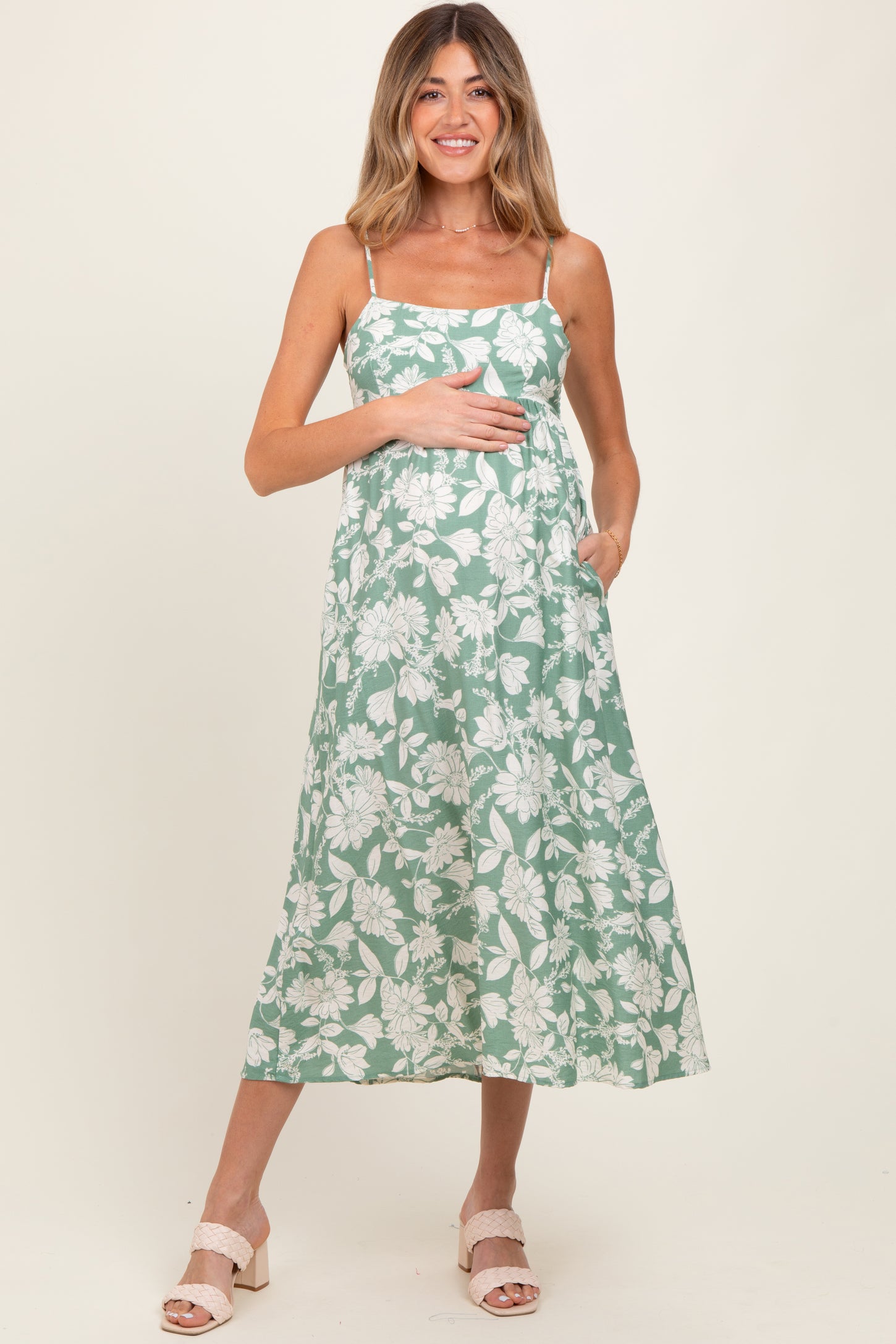 woman wearing a green floral maternity dress with adjustable straps, holding her baby bump and smiling