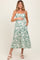 woman wearing a green floral maternity dress with adjustable straps, holding her baby bump and smiling
