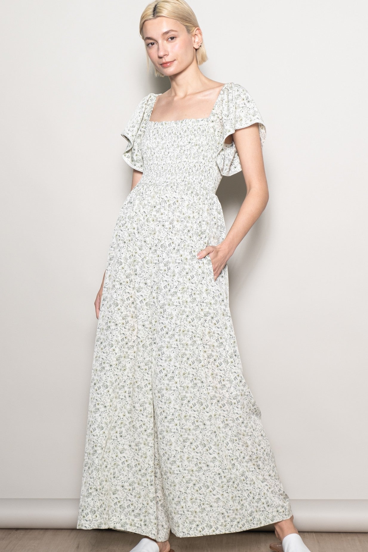 woman wearing a light floral print jumpsuit with short ruffle sleeves and a square neckline, standing with hands in pockets