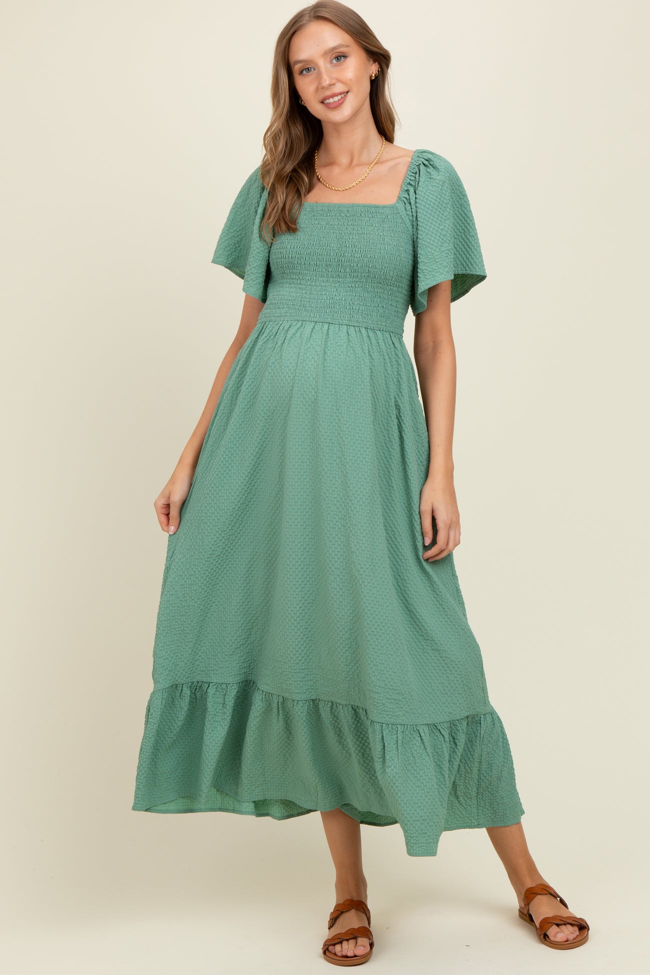 woman wearing a green maternity maxi dress with a square neckline and short puff sleeves, standing in a relaxed pose