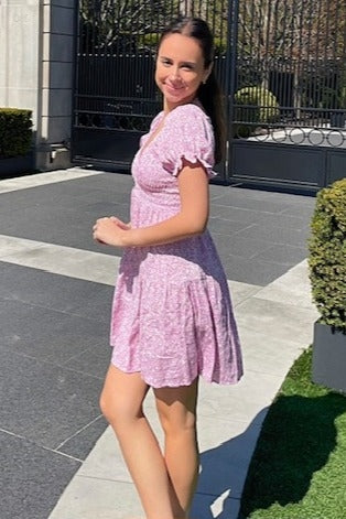 woman wearing a pink floral short sleeve dress, smiling and standing on a pathway outside