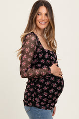woman wearing a black floral maternity top with sheer long sleeves and holding her baby bump