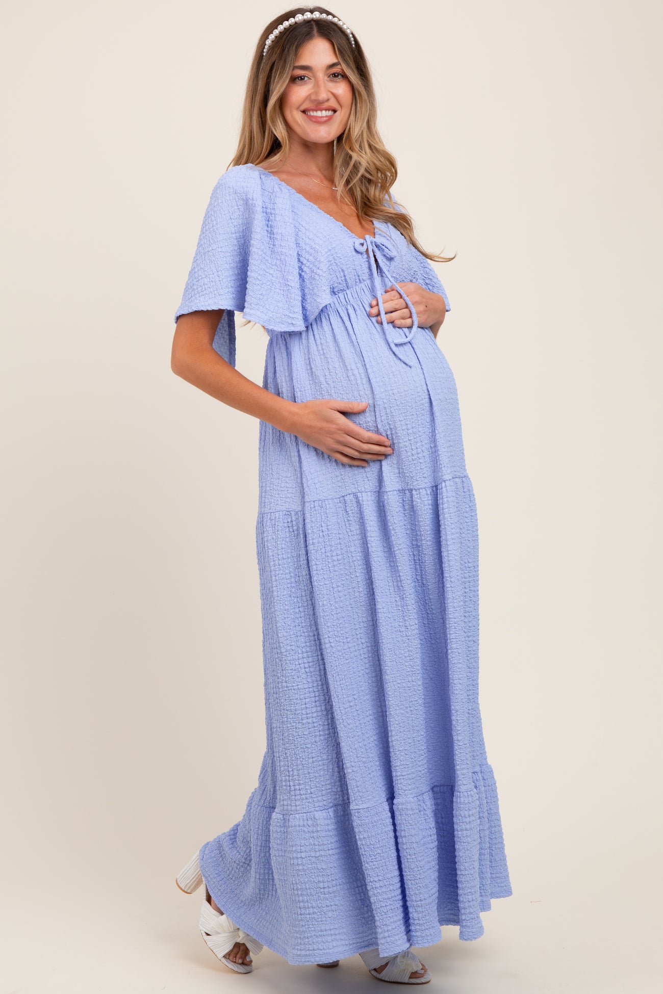 woman wearing a light blue maternity maxi dress with short sleeves and a tie detail, holding her baby bump