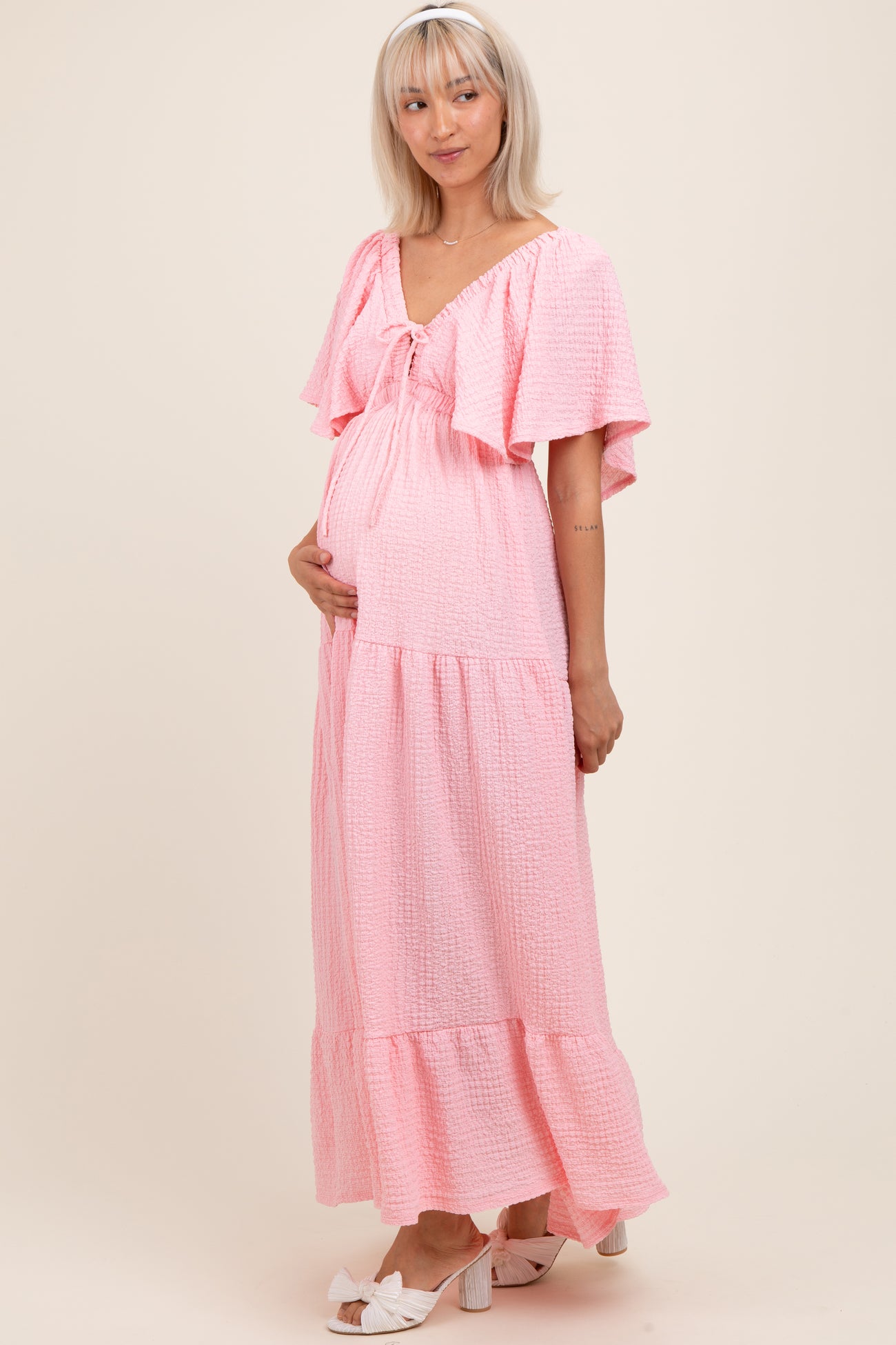 woman wearing a pink maternity maxi dress with frill sleeves and a tiered skirt, highlighting her baby bump