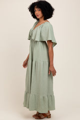 woman wearing a light green tiered maxi dress with a ruffled cape sleeve and a tie detail at the neckline