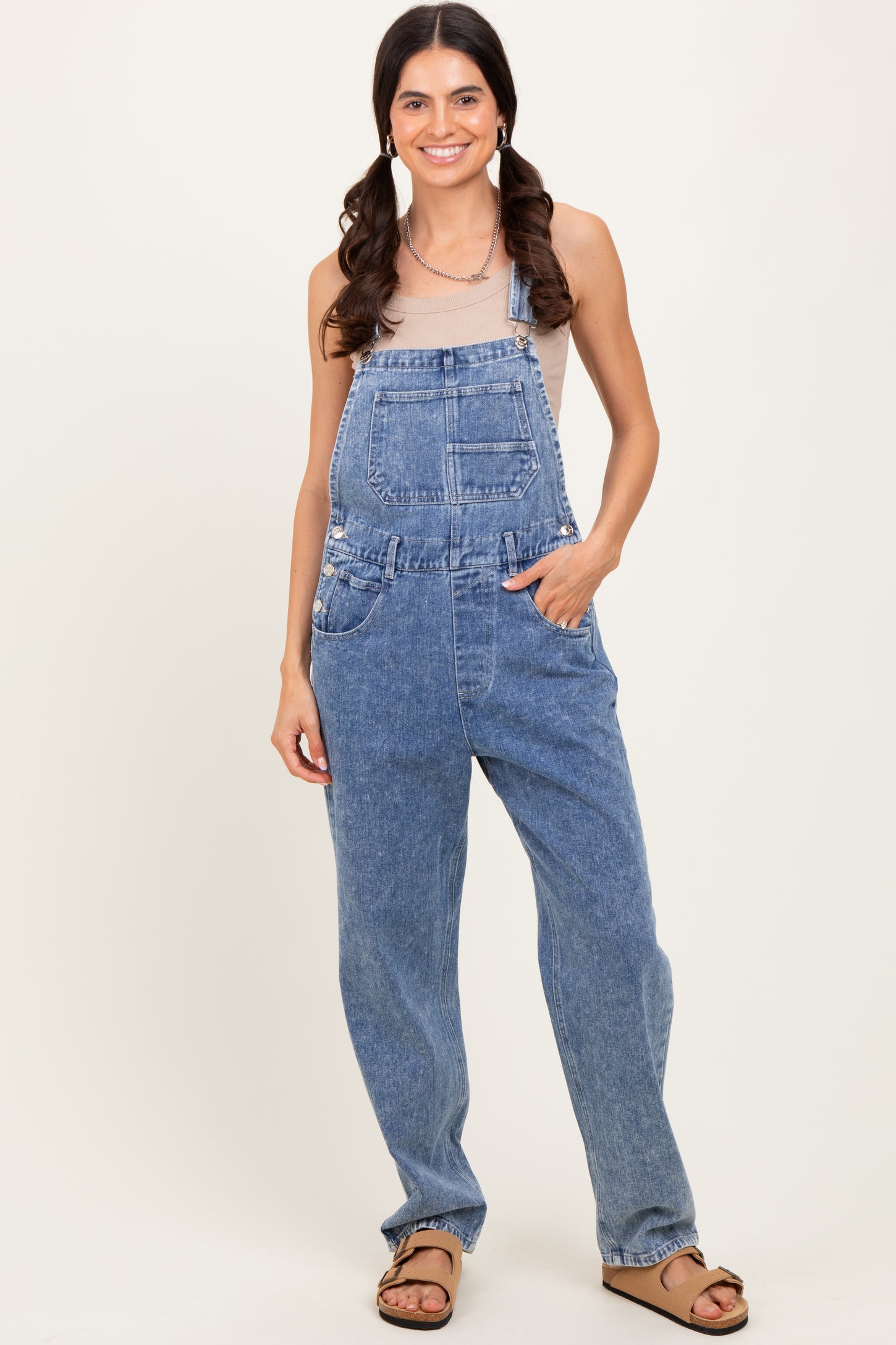 woman wearing light blue denim overalls with a fitted top and casual sandals, standing with hands in pockets