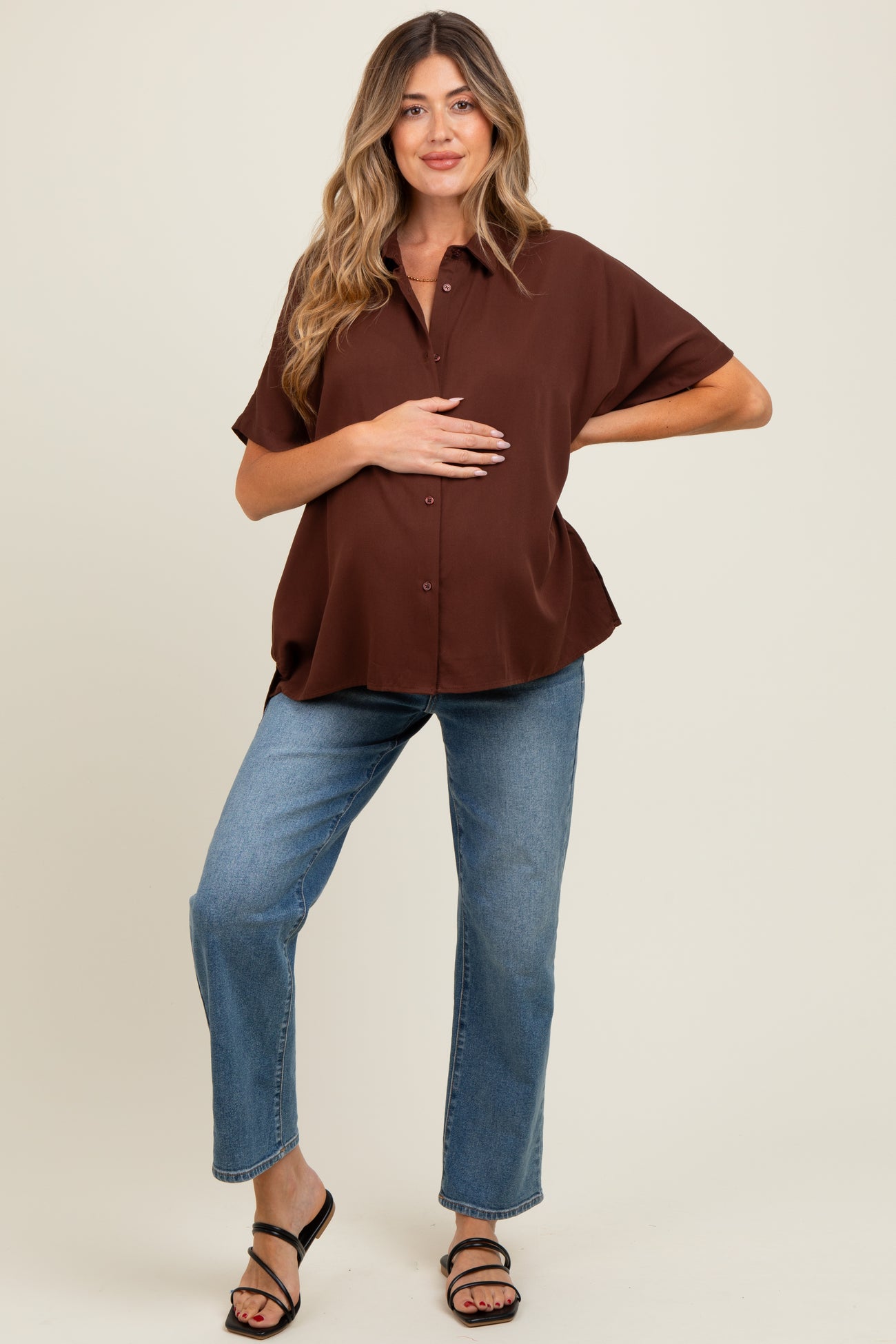 woman wearing a brown oversized maternity shirt and light blue jeans, holding her baby bump