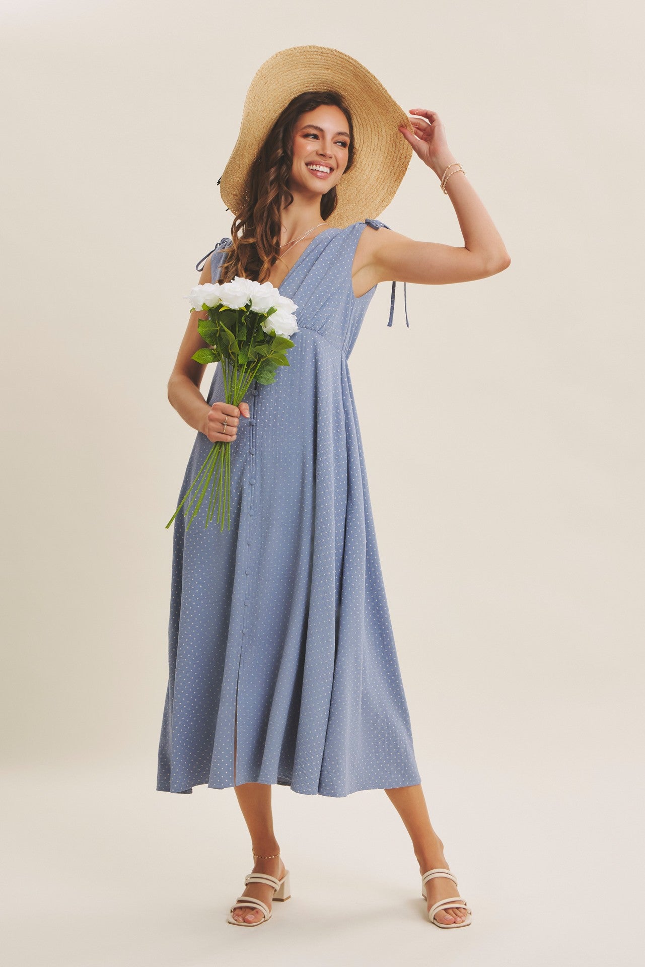 woman wearing a light blue sleeveless midi dress with tie straps, holding white flowers and wearing a large straw hat