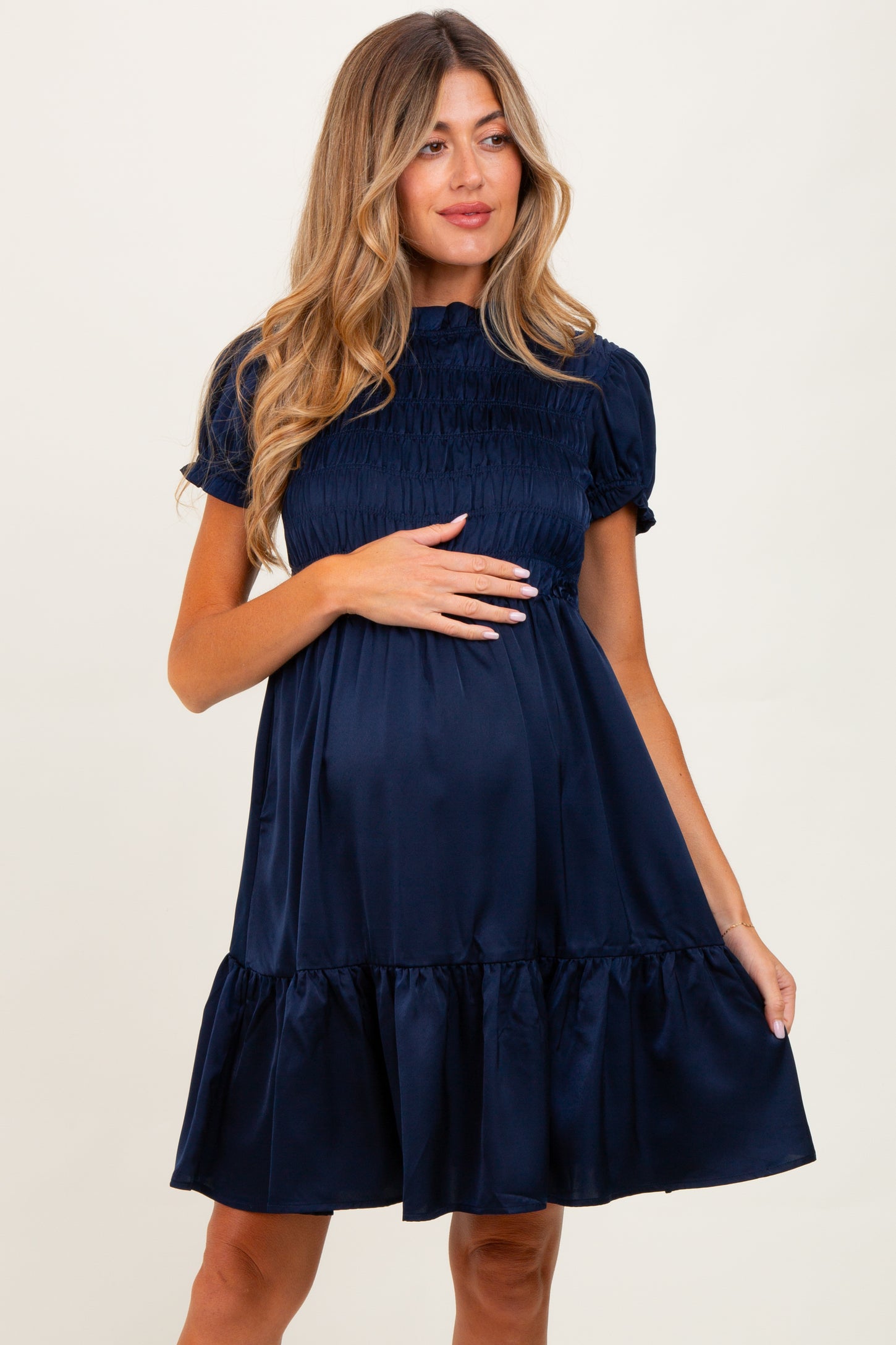 woman wearing a navy blue short sleeve maternity dress with smocked bodice and ruffled hem, holding her baby bump
