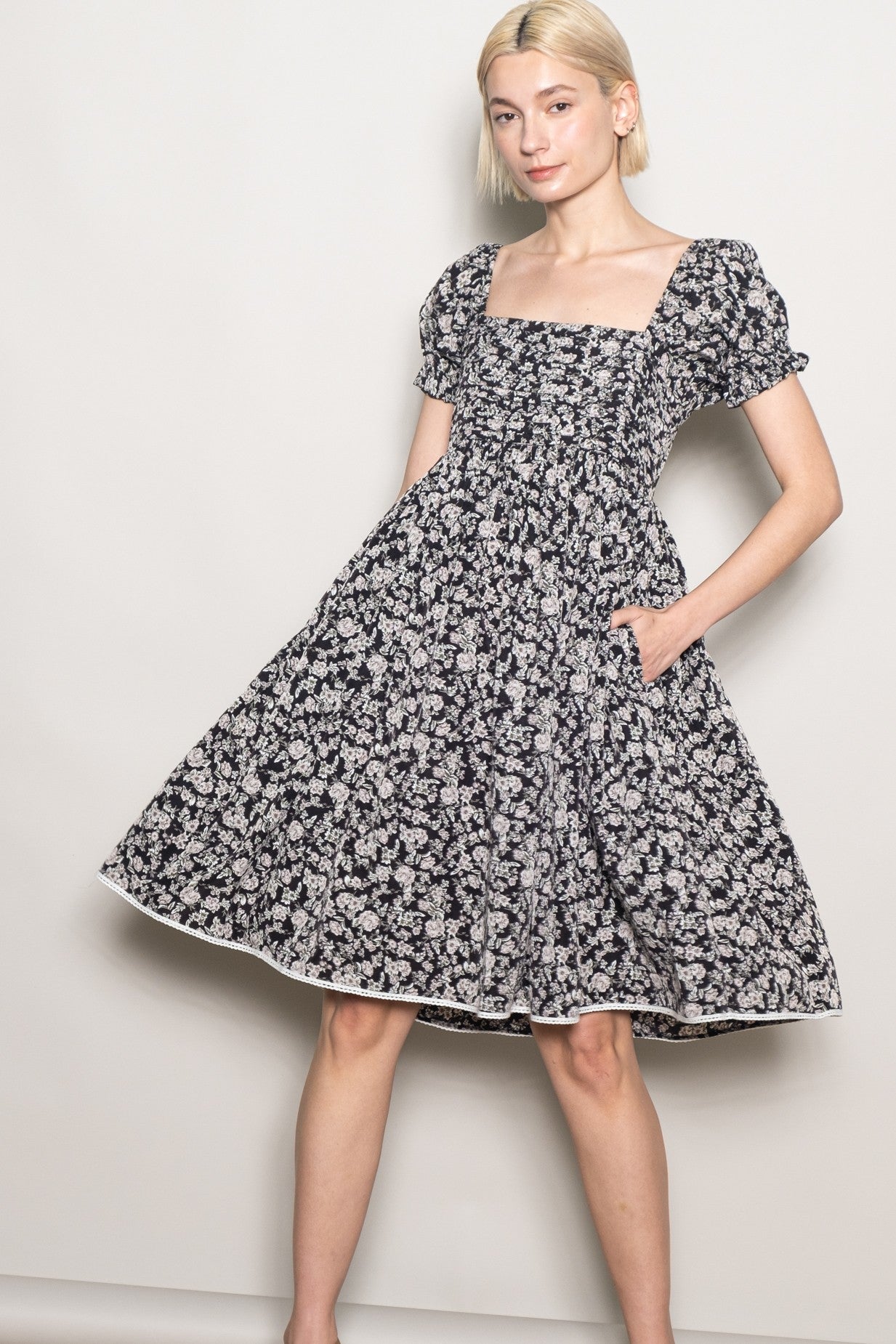 woman wearing a black floral puff sleeve dress with a square neckline and pockets, standing against a neutral background