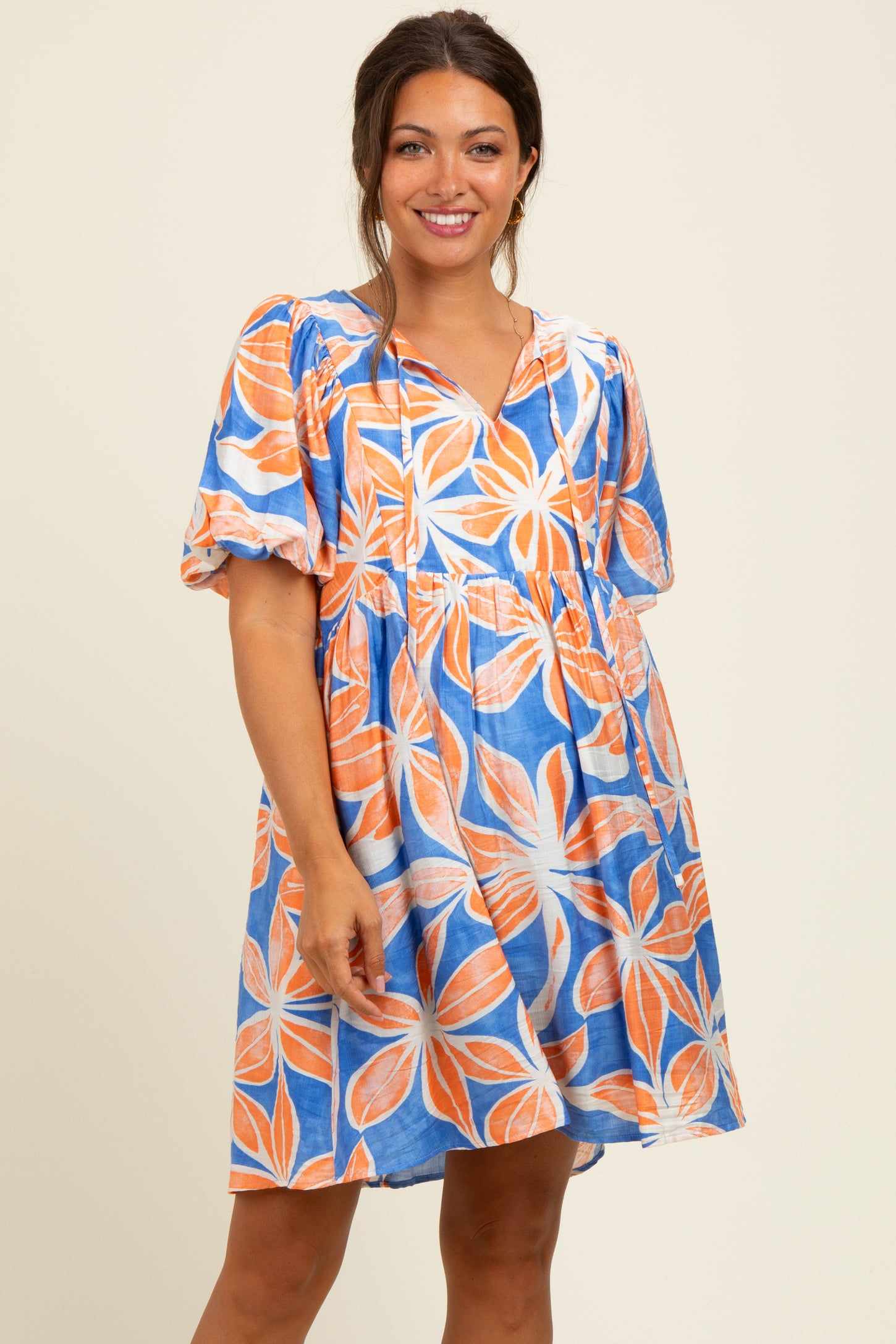 woman wearing a maternity short sleeve dress with a floral pattern in blue and orange, smiling and standing with hands on hips