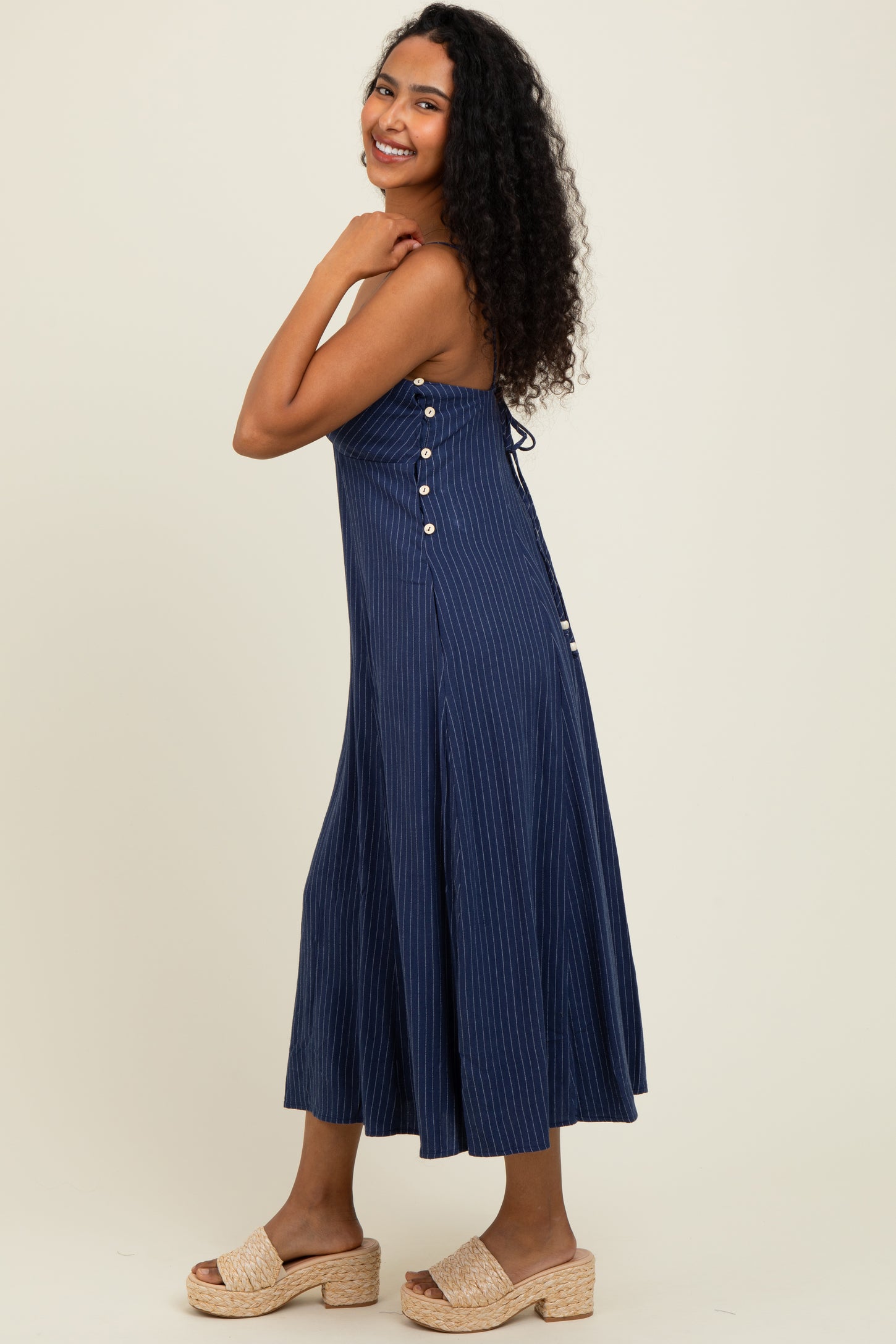 woman wearing a navy blue striped midi dress with button details and tied straps, paired with beige slip-on sandals