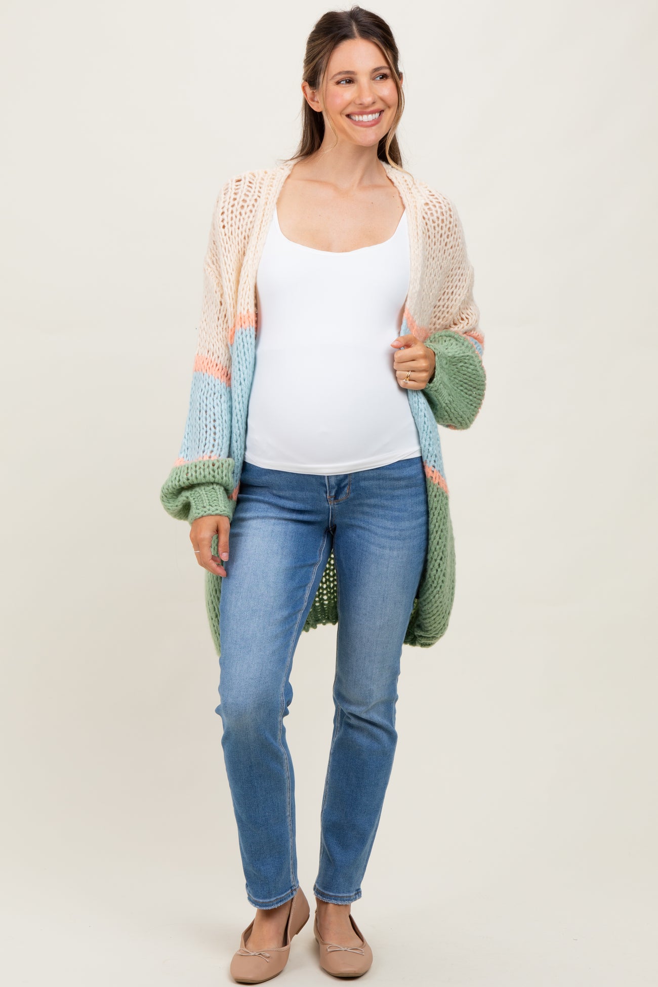 woman wearing a colorful open-knit maternity cardigan and a white tank top, showcasing her baby bump, paired with blue jeans