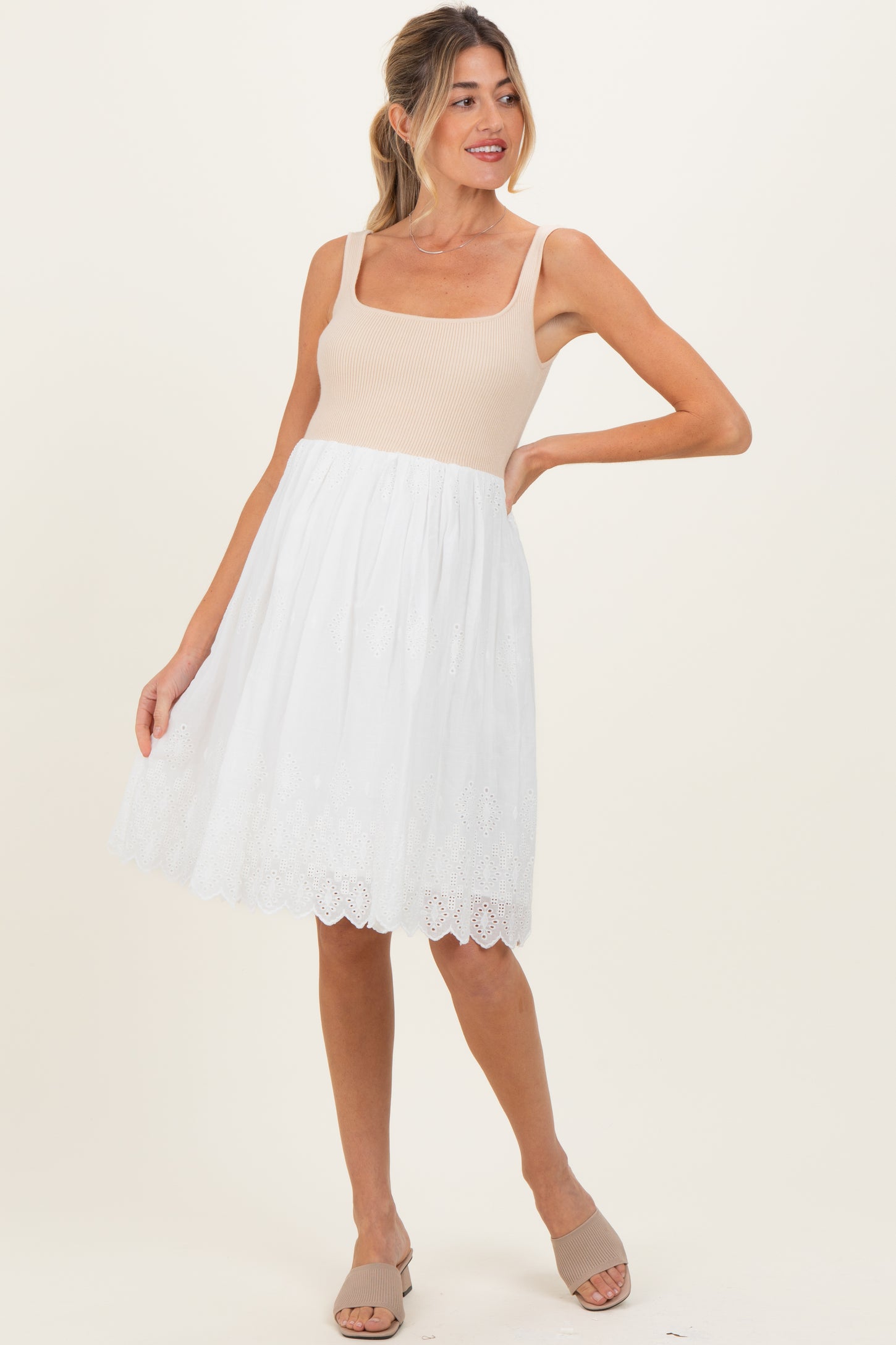 woman wearing a beige maternity dress with a fitted bodice and a white eyelet skirt, posing with one hand on her hip