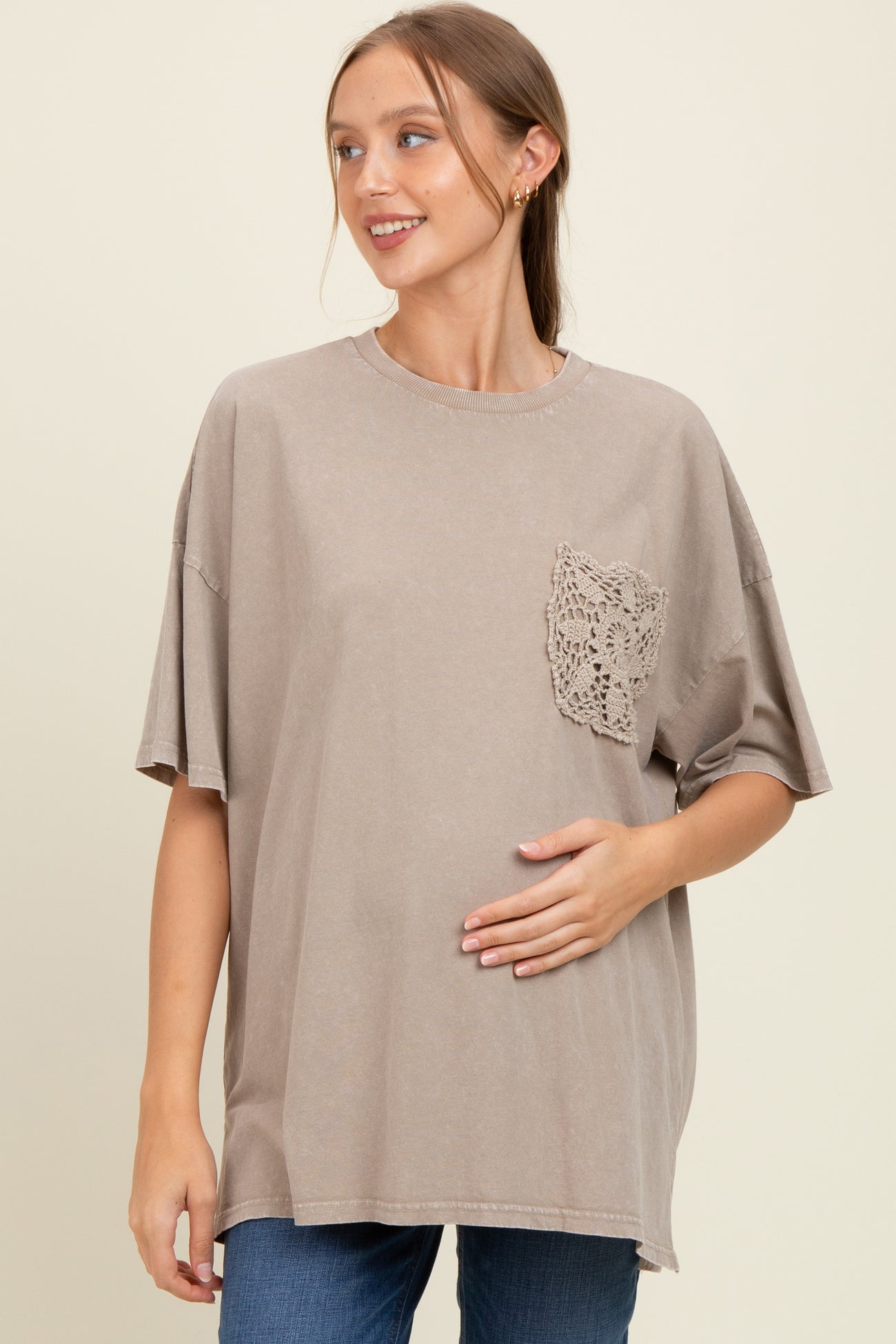woman wearing a taupe oversized maternity t-shirt with lace pocket, gently cradling her baby bump