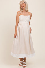 woman wearing a beige maternity dress with spaghetti straps and a flowy skirt, smiling and standing with hands in pockets