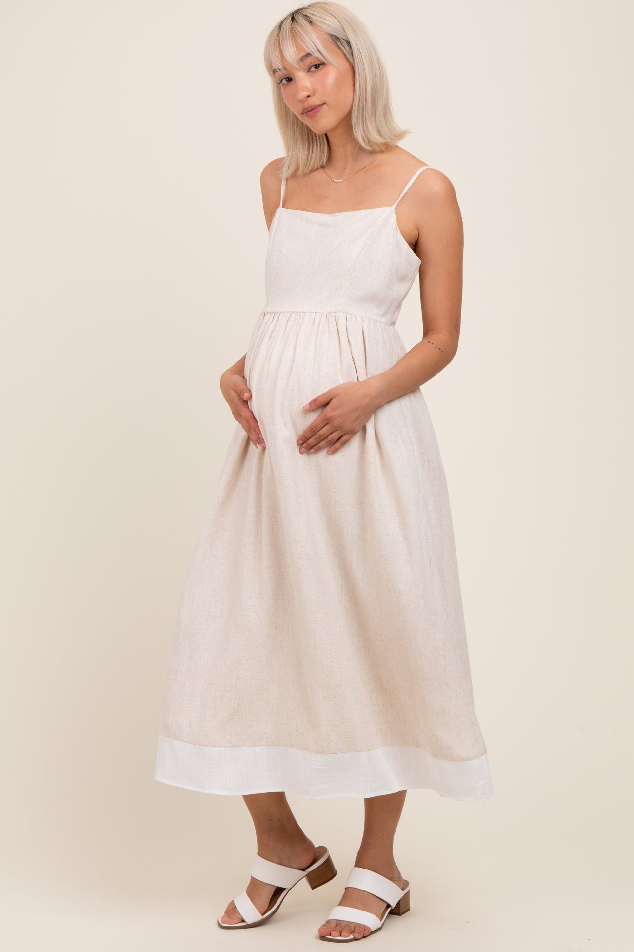 woman wearing a light beige maternity dress with spaghetti straps and a flowing skirt, holding her baby bump