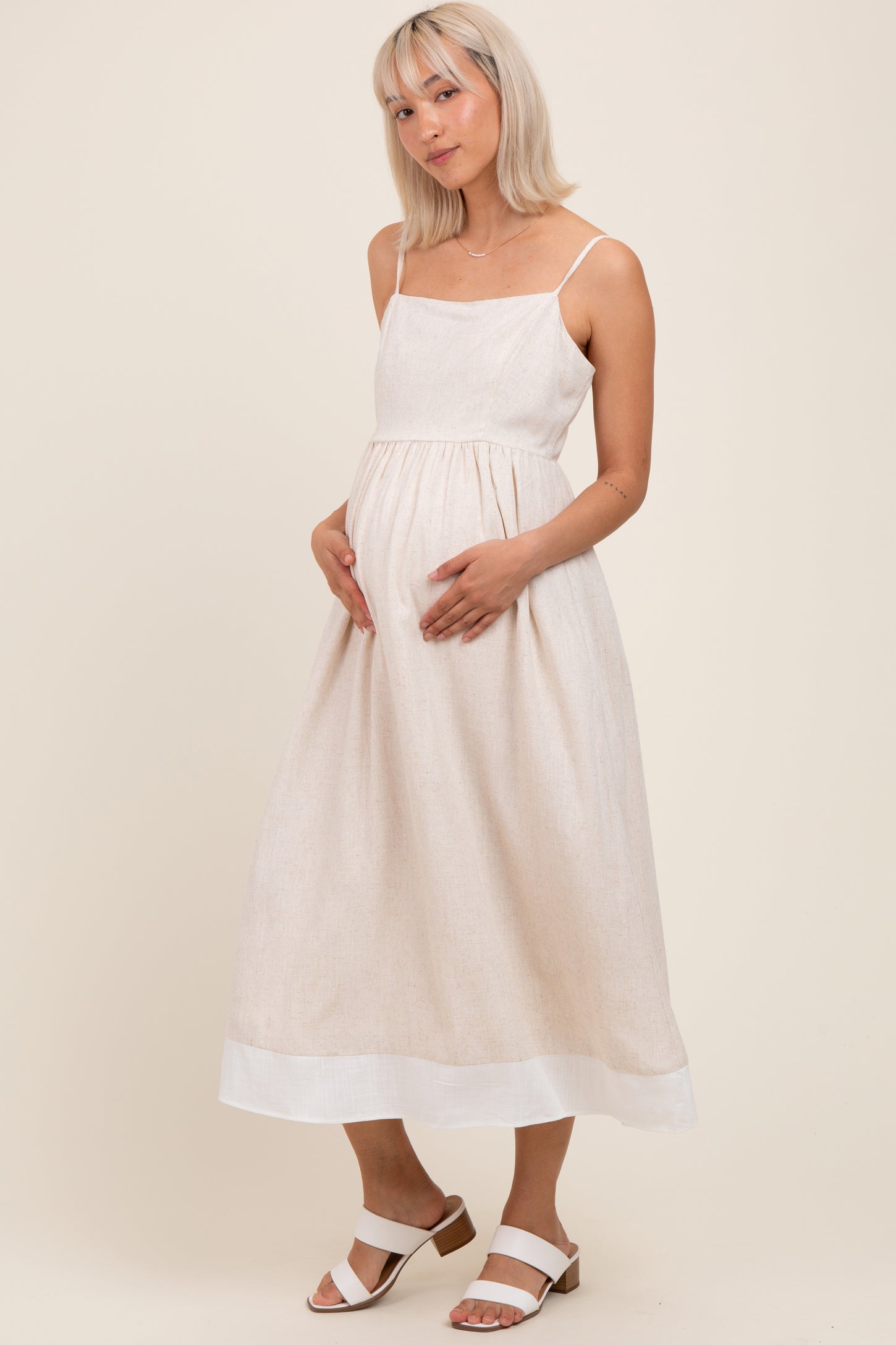 woman wearing a light beige maternity dress with spaghetti straps and a flowing skirt, holding her baby bump