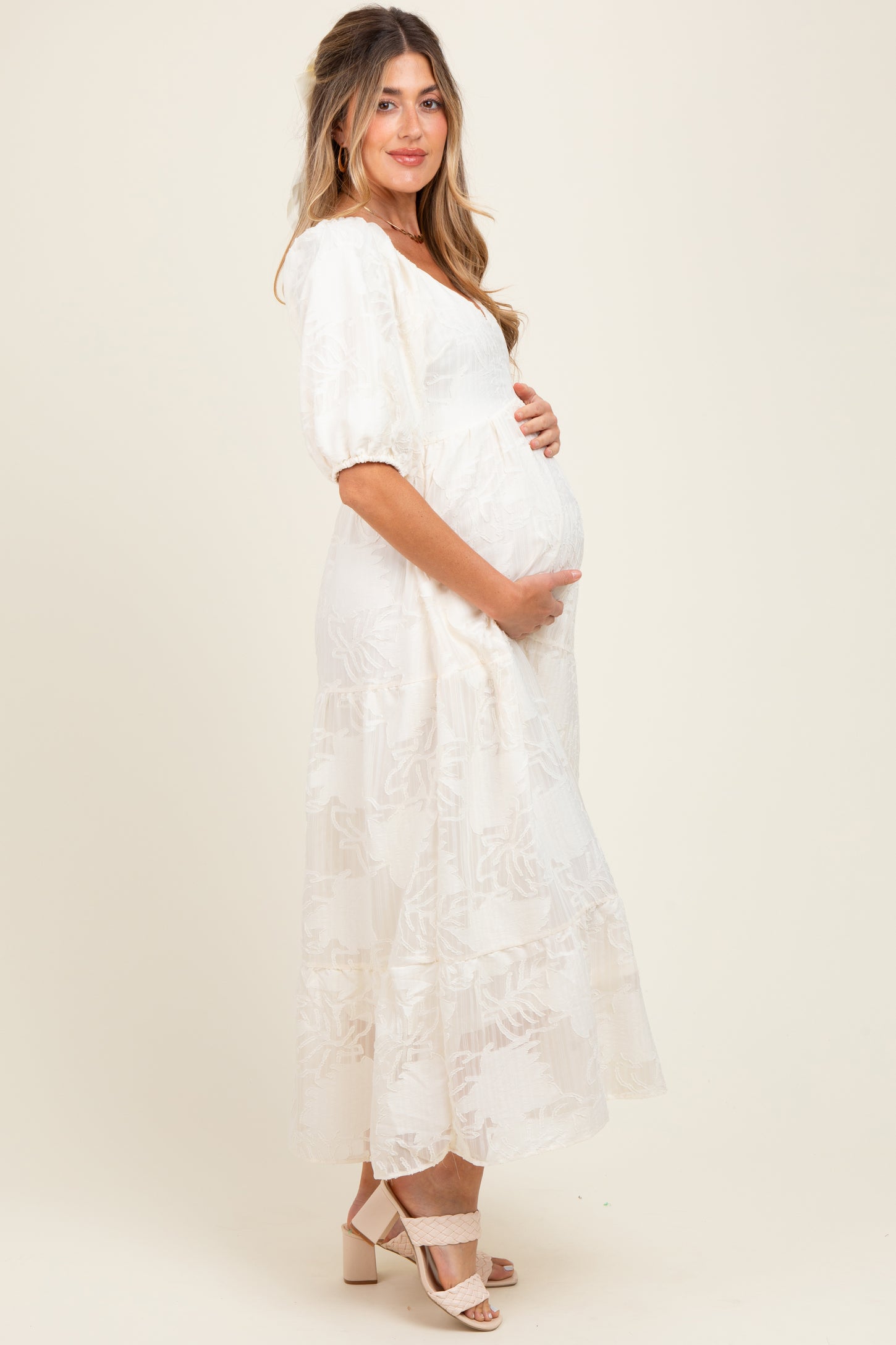 woman wearing a light beige maternity maxi dress with puff sleeves and tiered skirt, gently cradling her baby bump