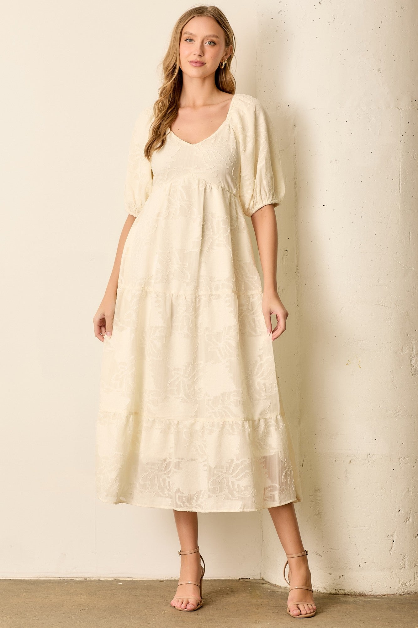 woman wearing a cream puff sleeve maxi dress with floral details and a v-neckline, standing against a textured wall