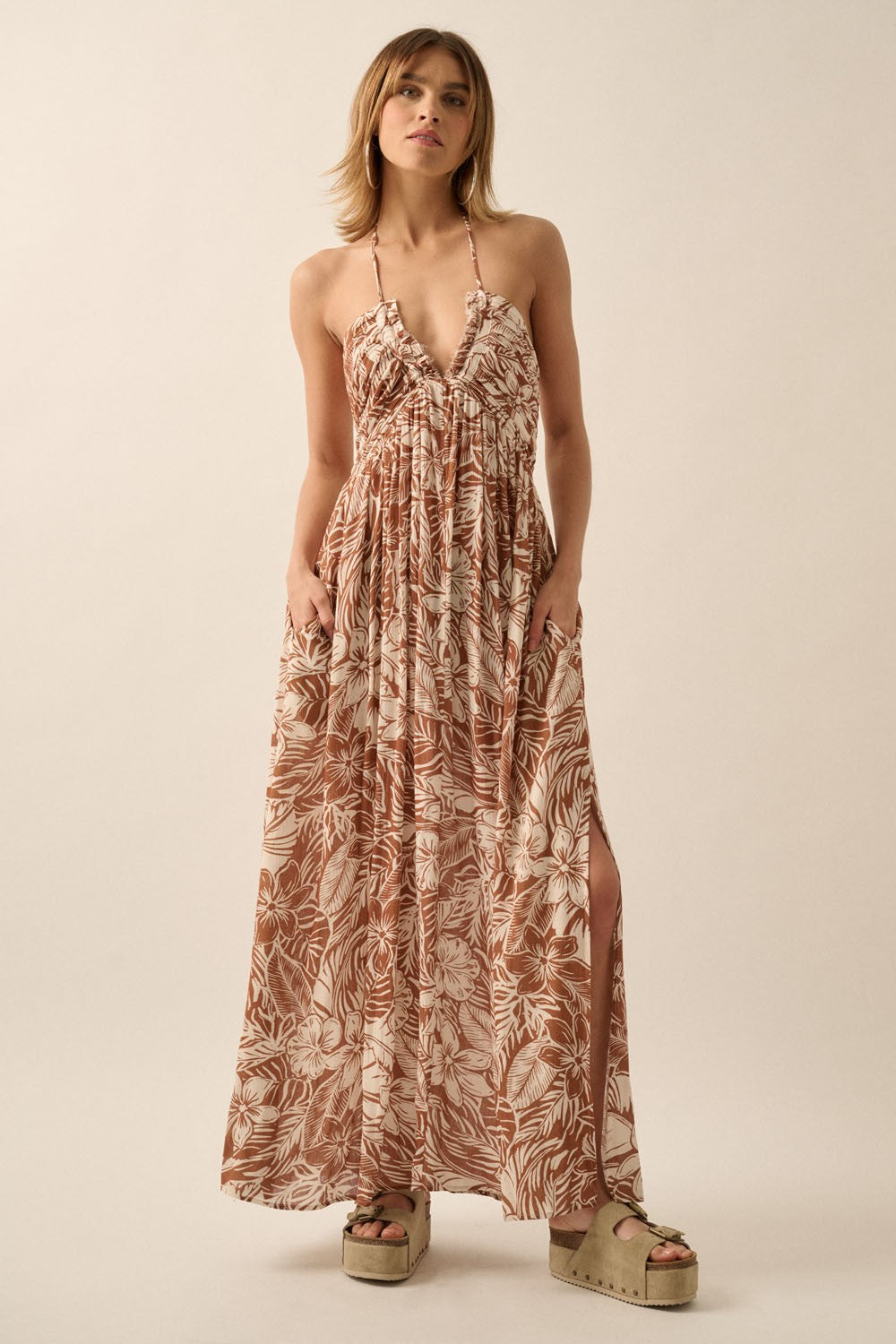 woman wearing a brown floral maxi dress with a deep v-neck and side slits, standing with hands in pockets