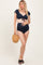 woman wearing a navy blue ruffled bikini top with tie detail and matching high-waisted bottoms, holding a straw bag, showing baby bump