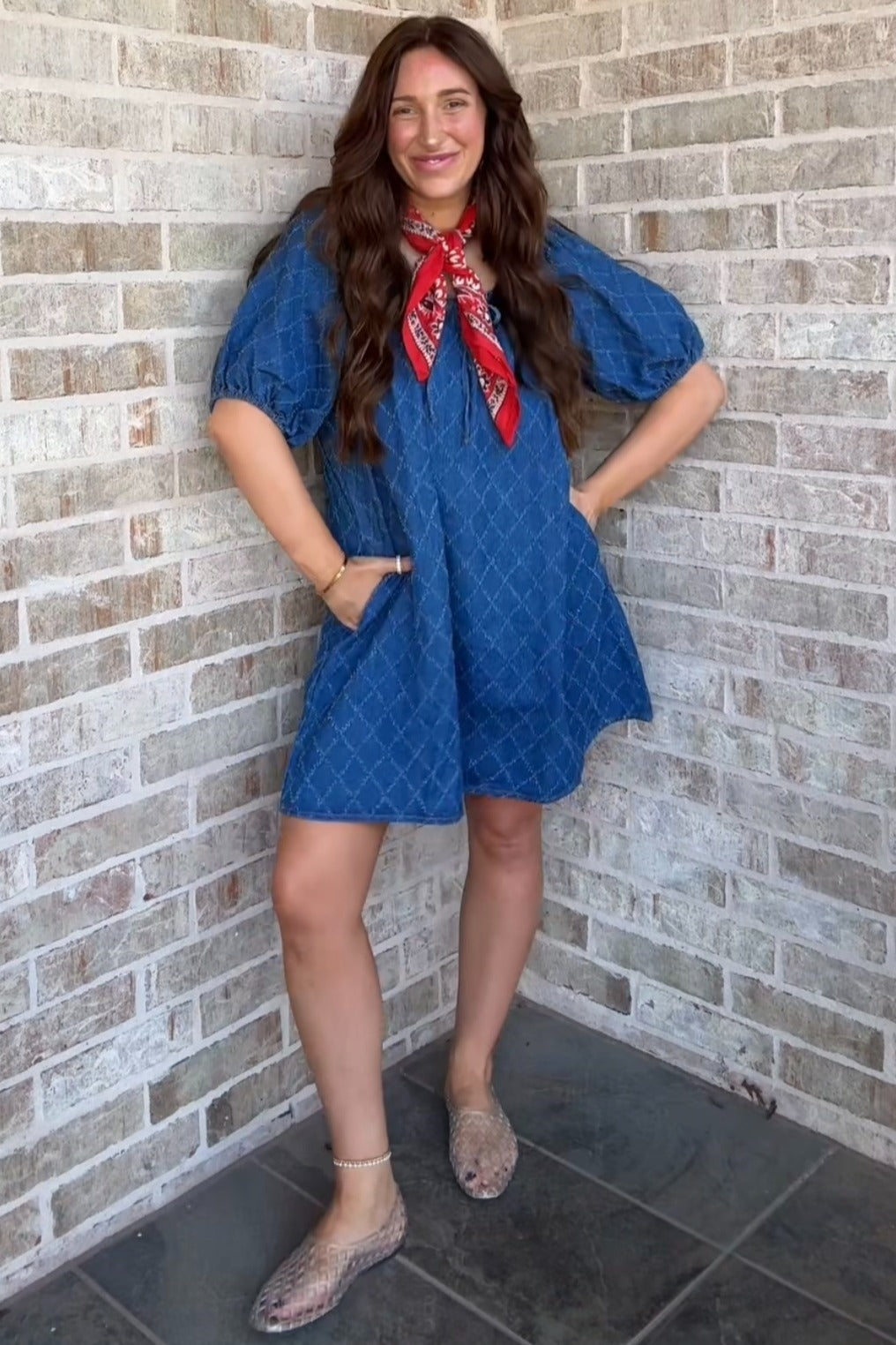 woman wearing a blue short patterned dress with puff sleeves and pockets, accessorized with a red scarf, standing against a brick wall