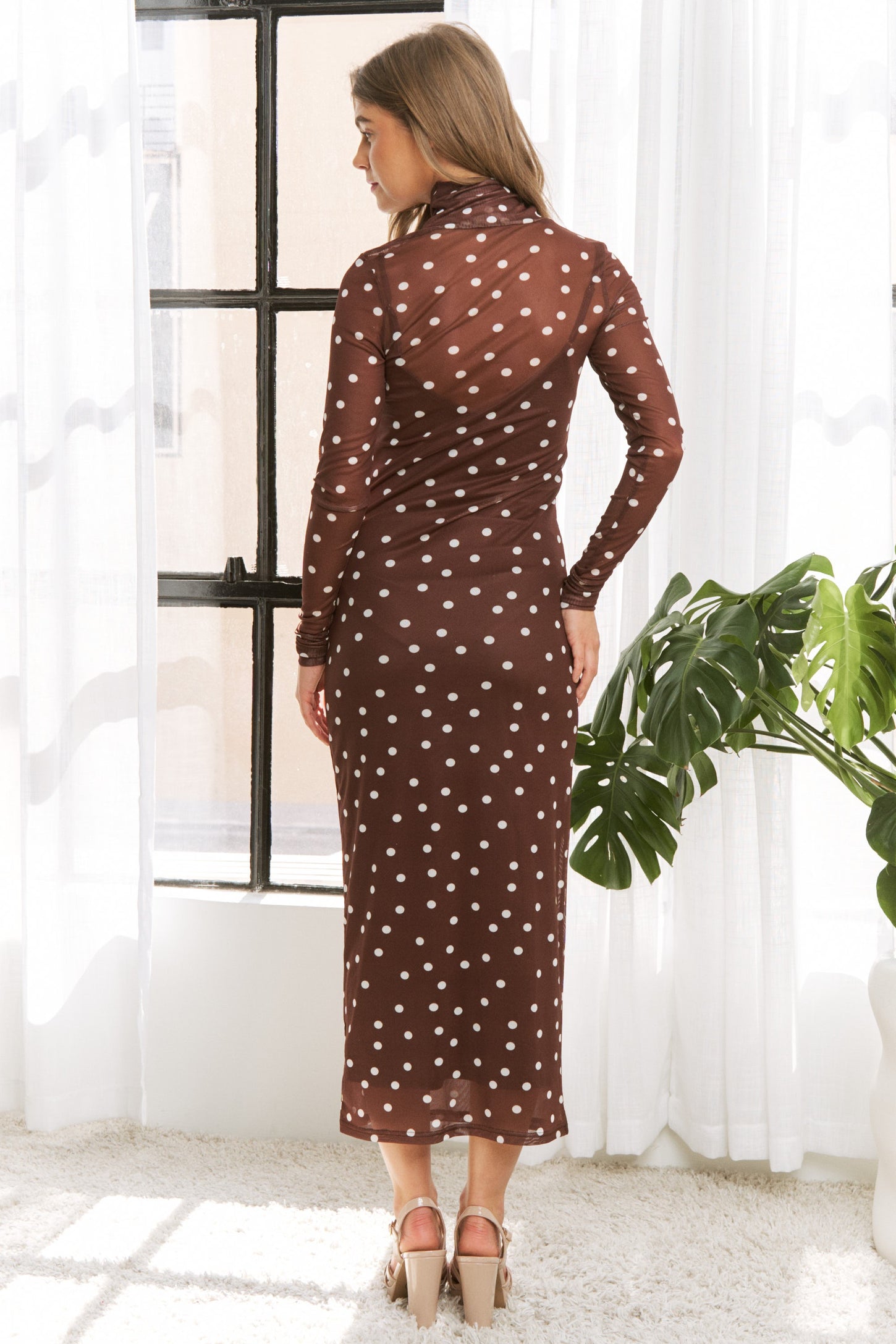 woman wearing a brown polka dot sheer long sleeve dress with a form-fitting silhouette, standing with her back to the camera