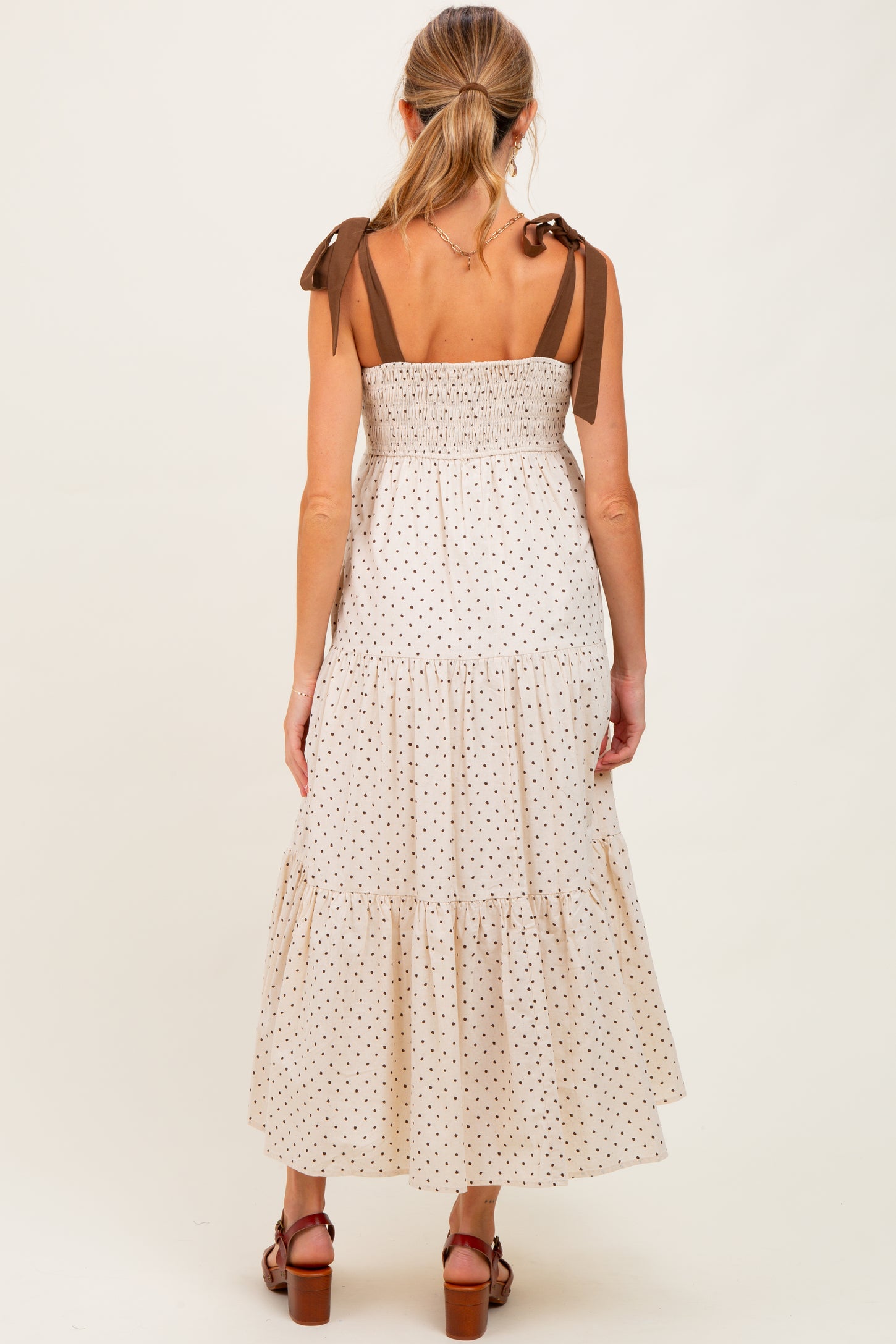 woman wearing a cream polka dot maternity maxi dress with brown tie straps and a smocked bodice, showing a baby bump from behind