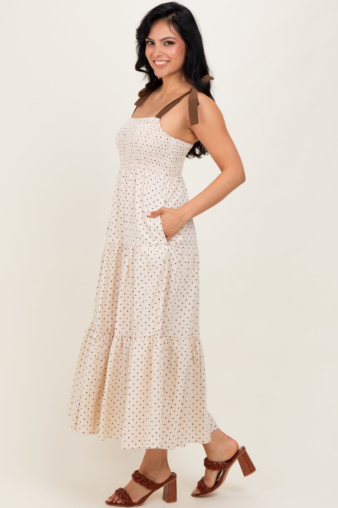 woman wearing a cream polka dot maxi dress with brown straps and pockets, styled with brown heeled sandals