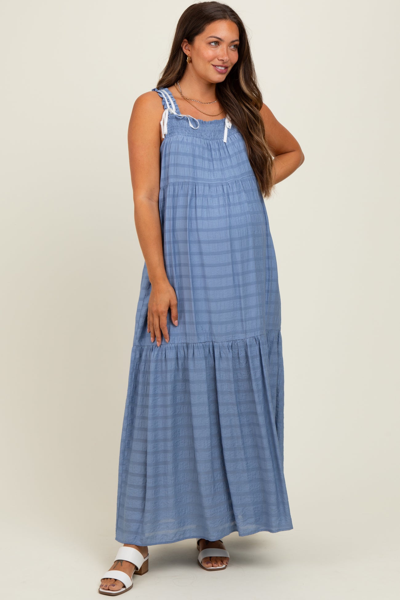 woman wearing a blue striped maternity maxi dress with adjustable straps and a tiered hem, posing with one hand on her hip