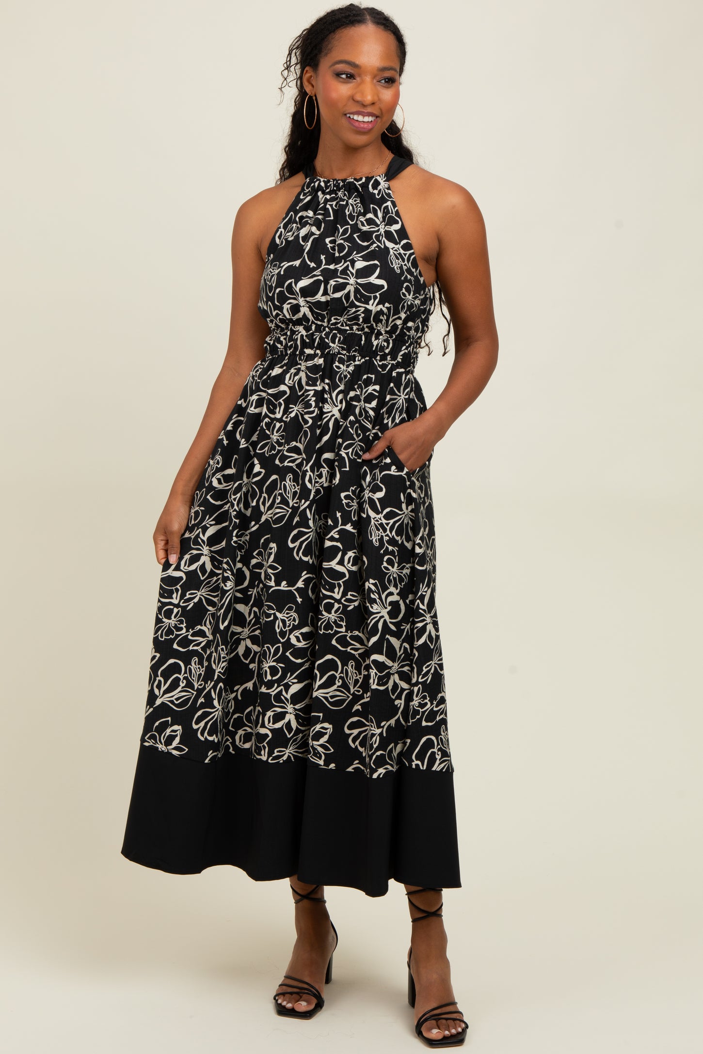 woman wearing a black halter neck maxi dress with white floral print and pockets, styled with strappy heels